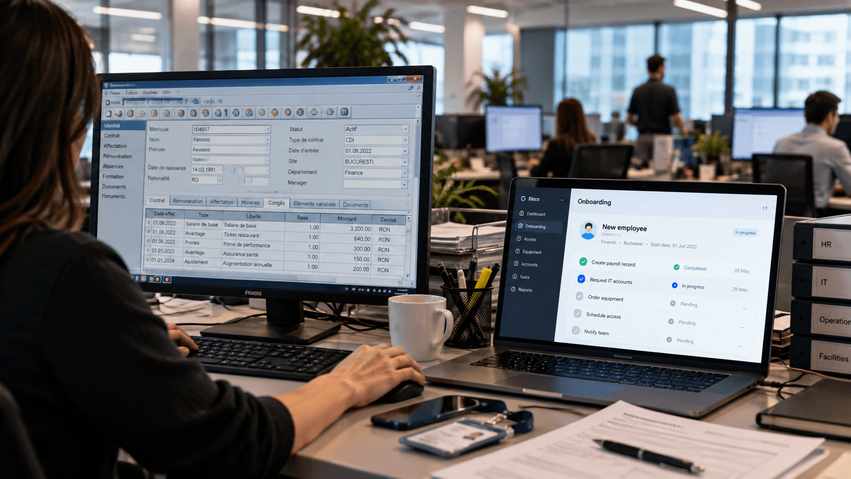 An office desk with an older desktop management application open on a large monitor beside a newer, more focused onboarding tool open on a laptop, with colleagues working at desks in a bright office in the background.